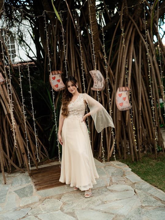 Retrato de corpo inteiro ao ar livre, com vestido claro, capa esvoaçante e maquiagem delicada.