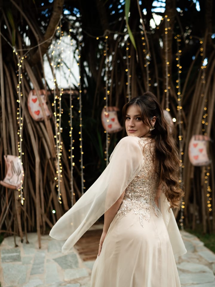 Ao ar livre, entre fios de luz, uma mulher de vestido claro olha por cima do ombro; pele iluminada e maquiagem suave.