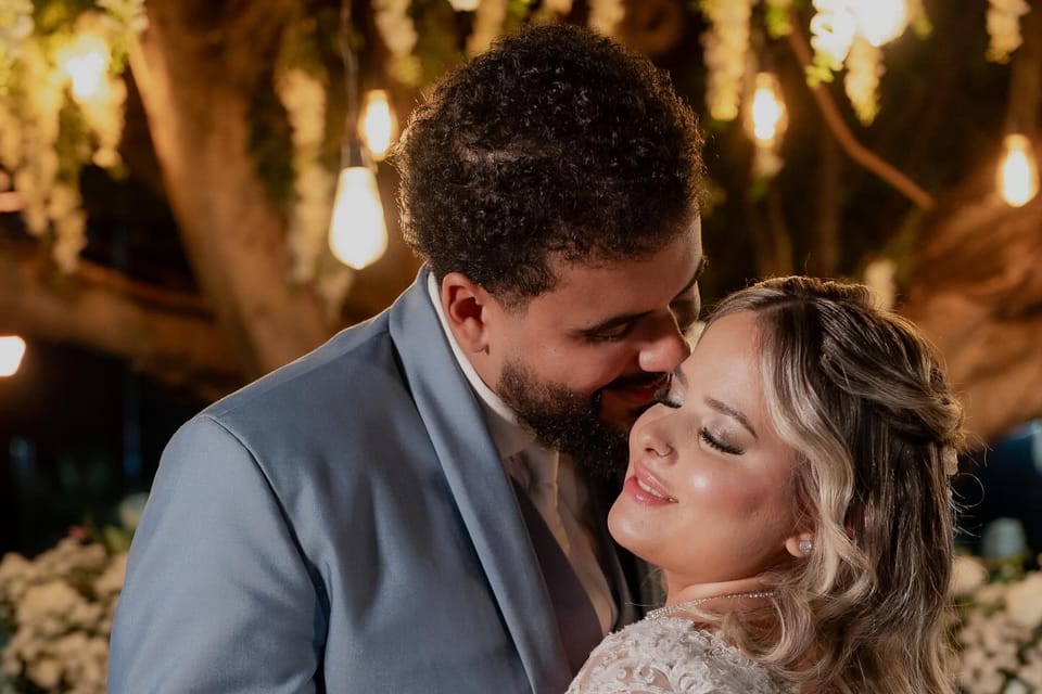 Em um casamento, a noiva sorri de olhos fechados enquanto recebe um beijo no rosto; pele iluminada e lábios com brilho.